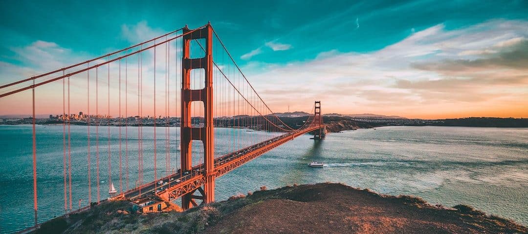 Golden Gate Bridge spans the blue bay at sunset with a vivid teal sky and orange-red towers transitioning to city skyline in the distance.