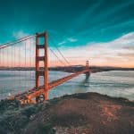 Golden Gate Bridge spans the blue bay at sunset with a vivid teal sky and orange-red towers transitioning to city skyline in the distance.