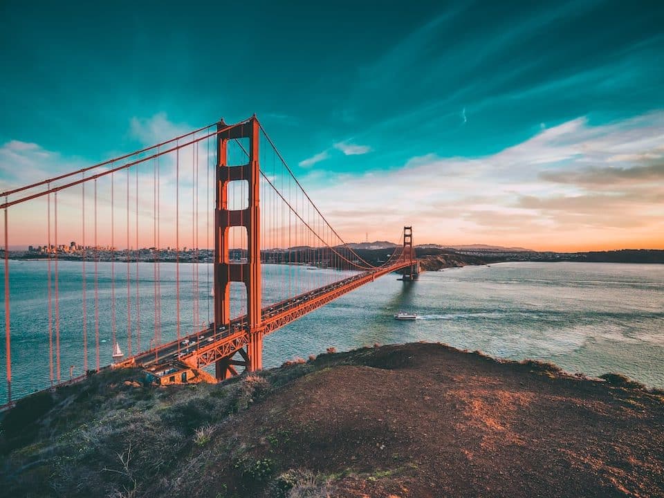 Golden Gate Bridge spans the blue bay at sunset with a vivid teal sky and orange-red towers transitioning to city skyline in the distance.